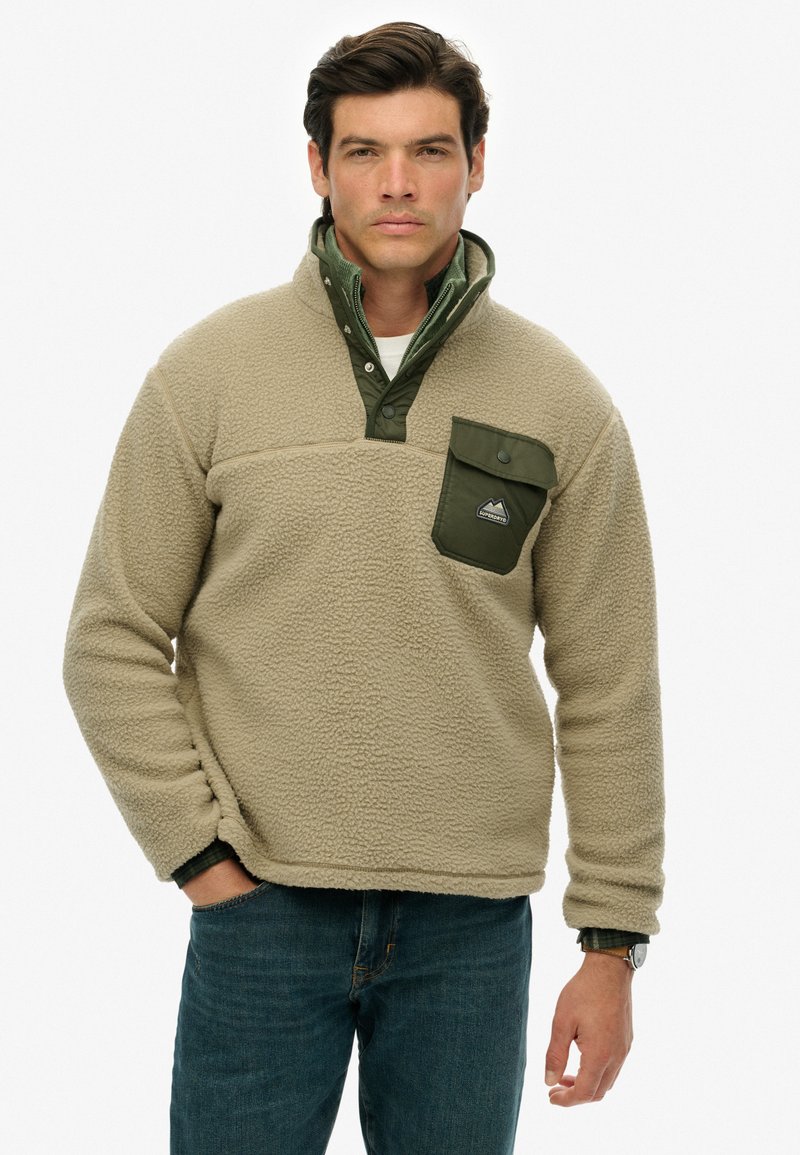 Beige fleece pullover med grøn krave og brystlomme, der har en tekstureret overflade og knaplukning. Elastiske manchetter og kant.