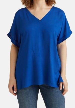 Kobieta nosząca luźną królewską niebieską bluzkę z krótkim rękawem i dekoltem w serek oraz niebieskie dżinsy, stojąca z ramionami swobodnie opuszczonymi wzdłuż ciała.