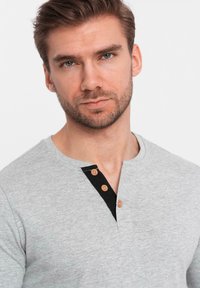 Närbild av en man med kort brunt hår, blå ögon och skägg som bär en grå henley-tröja med tre träknappar.