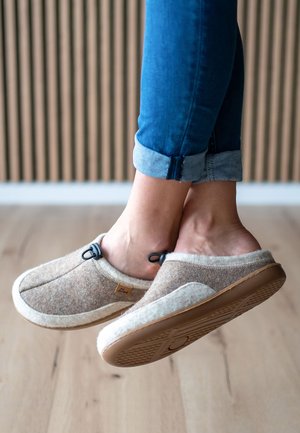 Chaussons en feutre brun avec une bordure en feutre blanche et une semelle en caoutchouc ; dotés d'un cordon élastique et d'un petit logo sur le côté.