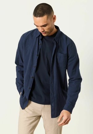 Mand iført åben mørkeblå skjorte med knapper over navy t-shirt og beige bukser, kigger ned med hænderne nær lommerne.