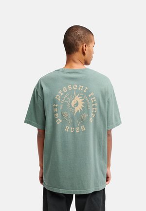 Uomo che indossa una t-shirt verde oversize con una grafica beige di sole, yin-yang, due mani e testo con la scritta "passato presente futuro, l’equilibrio degli opposti, RVCA."