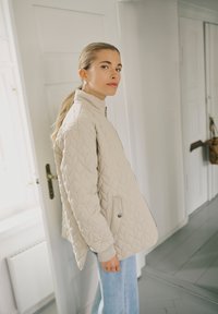 Heledate hobusesabaga naine, kes kannab beežikat püstlõiget jope ja helesiniseid teksapükse, asub siseruumides, valge ukse ja seina lähedal.