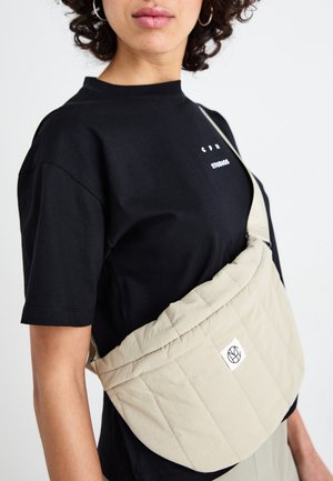 Sac à bandoulière beige matelassé avec une texture douce, arborant une petite étiquette logo et une bandoulière ajustable, porté sur un t-shirt noir.