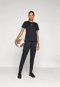 Črna majica s turkiznim logotipom, kombinirana s črnimi športnimi hlačami in črimi čevlji. V desni roki drži nogometno žogo.