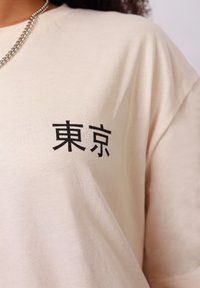 Άτομο που φοράει μπεζ πουκάμισο με μαύρους ιαπωνικούς χαρακτήρες για το «Τόκιο» στο στήθος και με ασημένιο κολιέ αλυσίδα ορατό.