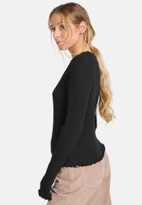 Schwarzes, geripptes Langarmoberteil mit rundem Ausschnitt und fransigem Saum. Kombiniert mit hellbeigen Cordhosen, die sichtbar Taschen haben.