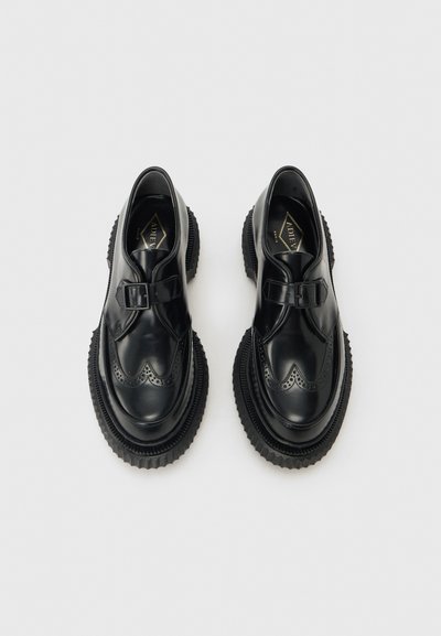 Ζεύγος μαύρα δερμάτινα παπούτσια monk strap με παχιές αυλακωτές σόλες και διακοσμητικές τρύπες brogue, τοποθετημένα δίπλα-δίπλα σε ανοιχτό φόντο.