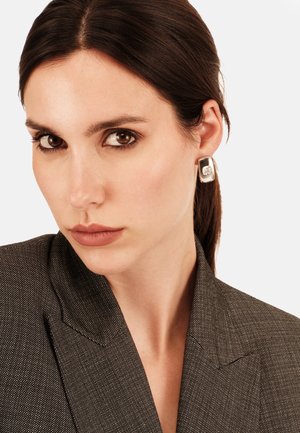 Pendientes rectangulares de plata con una superficie texturizada, llevados por una persona con un blazer oscuro y estampado. El fondo es blanco liso.