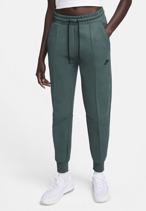 Grønne Nike træningsbukser med snøre i taljen, sidelommer og tilspidsede ben med en sort logo på venstre lår. Blødt stof.