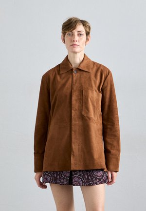Persona con capelli corti che indossa una giacca di suede marrone sopra una gonna a fantasia, in piedi contro uno sfondo grigio chiaro uniforme.