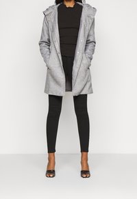 Mujer con un abrigo gris con capucha sobre un suéter negro de canalé, pantalones skinny negros y tacones abiertos negros, de pie contra un fondo blanco.