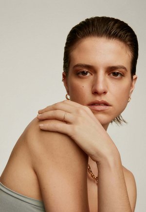 Persona joven con el cabello peinado hacia atrás, apoyando la mano con un delicado anillo sobre el hombro desnudo, lleva pequeños pendientes de aro y maquillaje neutro.