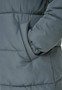 Veste grise rembourrée avec une poche latérale, dotée d'une texture lisse et de détails cousus, mettant en avant un design moderne.