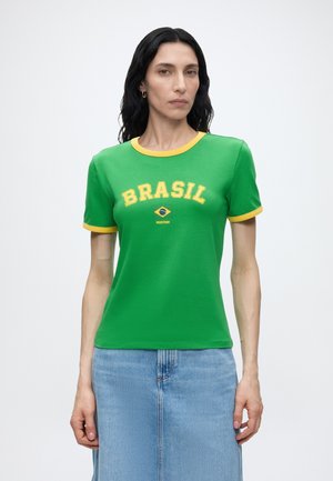 Mujer con largo cabello negro que lleva una camiseta verde con ribete amarillo y el texto "BRASIL" sobre una pequeña bandera de Brasil, combinada con una falda vaquera azul.
