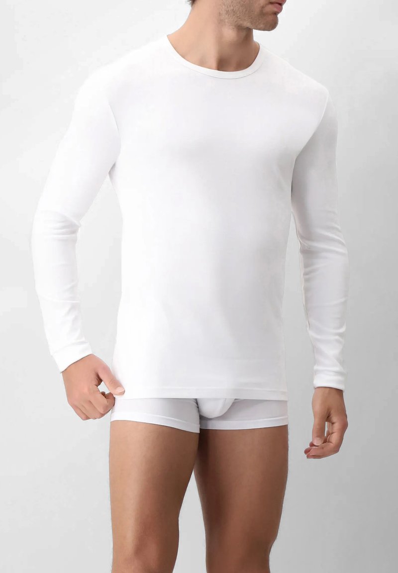 Uomo che indossa una camicia a maniche lunghe aderente bianca e boxer bianchi, in piedi contro uno sfondo chiaro e semplice.