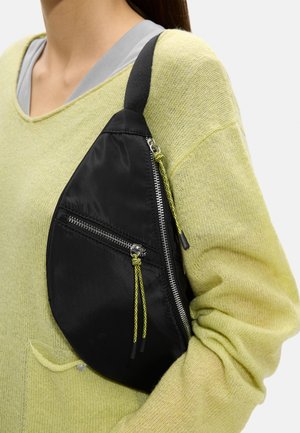 Czarna torebka na ramię w kształcie kropli z srebrnymi zamkami i żółto-czarnymi sznurkami, noszona na bladożółtym dzianinowym swetrze.