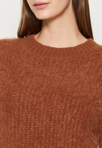 Brązowy sweter dziany o miękkiej, puszystej teksturze. Posiada elegancki okrągły dekolt oraz subtelne pionowe linie w wzorze materiału.