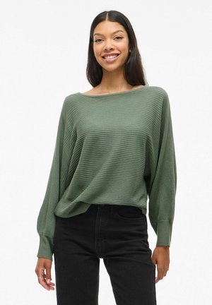 Pull en maille côtelée olive avec un col large et des manches longues, associé à un jean noir taille haute. Texture subtile et coupe décontractée.