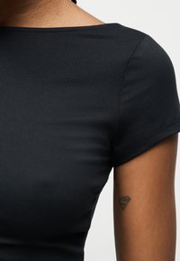 Κοντινό πλάνο ενός ατόμου που φοράει στενό μαύρο κοντομάνικο πουκάμισο, δείχνοντας τον ώμο, το πάνω μέρος του βραχίονα και ένα μικρό τατουάζ στον βραχίονα.