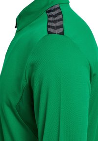 Grøn sportstrøje med et tekstureret, åndbart stof. Har sort mønstret accent på skulderen og en kvart lynlås ved halsen.
