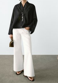 Black, pleated, oversized shirt with button closure; paired with white flared trousers and black flip-flops. Accessories include a small bag.