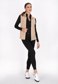 Beige ärmellose Weste mit hohem Kragen und frontalem Reißverschluss, getragen über einem schwarzen langärmligen Oberteil und schwarzen Leggings, kombiniert mit weißen Sneakers.