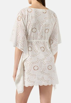 Vrouw die een witte eyelet kanten jurk draagt met bloemen uitgesneden patronen, losse mouwen en een golvende zoom, gezien van achteren.