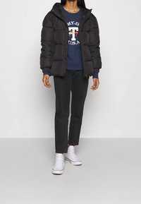 Czarna kurtka puchowa z kapturem, noszona na niebieskiej bluzie z graficznym logo, w połączeniu z ciemnymi dżinsami i białymi sneakersami.