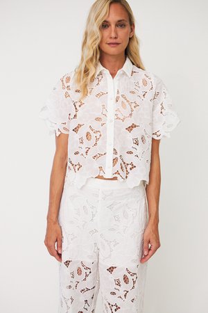 Femme portant un top boutonné en dentelle blanche à motifs floraux, manches courtes, et un pantalon en dentelle assorti à taille haute, debout devant un fond uni.