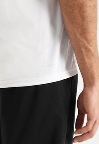 Camiseta atlética blanca Climalite con una textura suave, presenta una etiqueta en el dobladillo, combinada con pantalones cortos negros. La camiseta está diseñada para absorber la humedad.