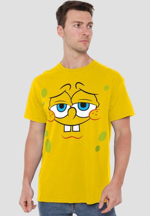 Gelbes Baumwoll-T-Shirt mit dem Gesicht eines Cartoon-Charakters, blauen Augen, ausgeprägten Zähnen und grünen Punkten, die auf dem Stoff aufgedruckt sind.