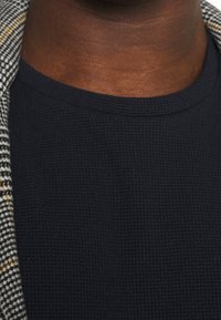 Schwarzes, strukturiertes T-Shirt mit rundem Ausschnitt, kombiniert mit einer gemusterten Jacke, die graue und gelbe Akzente am Kragen aufweist.