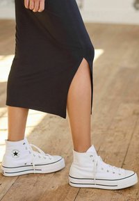 Baskets montantes blanches en toile avec des accents noirs, portées avec une robe noire à fente sur le côté. Texture lisse et design classique.