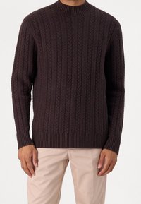 Pull en maille côtelée brun foncé avec un col rond, présentant un motif texturé et des poignets et un ourlet ajustés. Porté avec un pantalon beige clair.