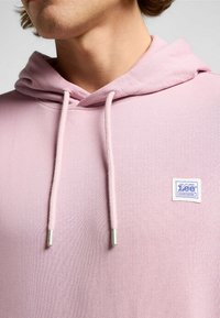 Jasnoróżowa bluza z kapturem z kieszenią typu kangur, sznurkami do regulacji i srebrnymi metalowymi końcówkami. Posiada małą niebiesko-białą metkę z logo marki.