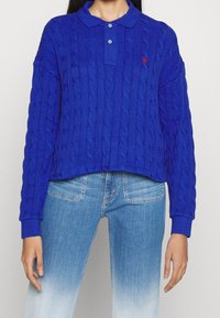 Blå kabelstickad kort tröja med polo-krage, som har tre knappar och ribbade manschetter. Matchad med ljusblå denimjeans.