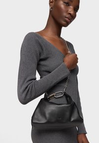 Mujer con vestido ajustado gris acanalado, llevando un bolso de cuero negro con correa de cadena plateada y broche metálico grande al hombro, sobre fondo blanco.