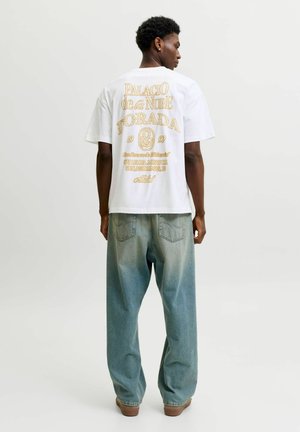 Jeune homme de dos portant un t-shirt blanc avec un texte jaune et un jean bleu délavé ample sur un fond uni.
