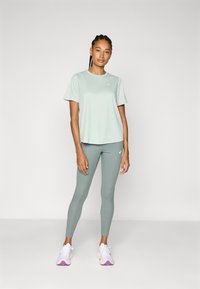 Ljusgrön kortärmad träningströja som matchas med leggings. Båda plaggen har logotypdetaljer. Modellen bär vita och rosa sneakers.
