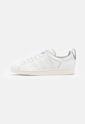 Baskets basses blanches Adidas Superstar avec côtés texturés, laçage à l'avant, bout coquillage et languette de talon grise sur fond blanc.