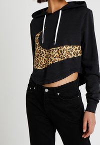 Person som bär en svart cropped hoodie med vita dragsnöre och en stor leopardmönstrad Nike-swoosh, tillsammans med högmidjade svarta jeans.