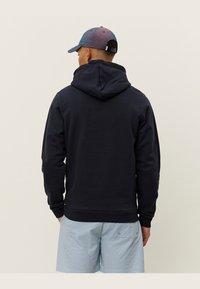 Dunkelblauer Kapuzenpullover aus weichem Stoff, mit langen Ärmeln, lockerer Passform und gerippten Bündchen. Getragen mit hellblauen gemusterten Shorts.