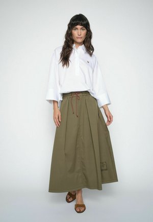 Mujer con una camisa blanca de cuello suelta metida en una falda larga verde oliva de talle alto con cordón delantero y sandalias marrones.