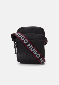 Borsa a tracolla nera con chiusura a zip, design imbottito e tracolla regolabile con testo ripetuto "HUGO" in bianco e rosso.