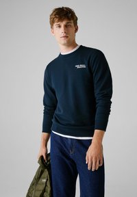 Marineblaue Sweatshirt aus weichem Material, mit gerippten Bündchen und Kragen. Weißes "RED BULL RACING" Logo auf der linken Brust. Das Model hält eine grüne Jacke.