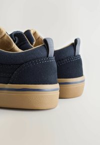 Chaussures en toile bleu marine avec accents marron, semelle texturée et détails de couture grise. Présente des petites boucles au talon pour un enfilage facile.