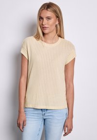 Kratká rukávová, světle žlutá ribová pletená blůza s kulatým výstřihem a volným střihem. Má horizontální pruhy a hladkou texturu.