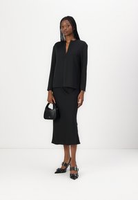 Top nero a maniche lunghe con scollo a V, abbinato a una gonna nera aderente. Gli accessori includono una piccola borsa nera a tracolla e tacchi neri con cinturini.