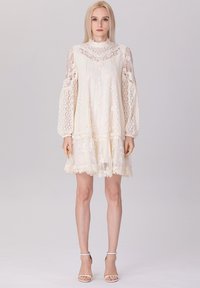 Robe en dentelle crème avec de longues manches bouffantes transparentes, broderies florales, col montant et ourlet évasé, complétée par des escarpins blancs à brides au niveau de la cheville.